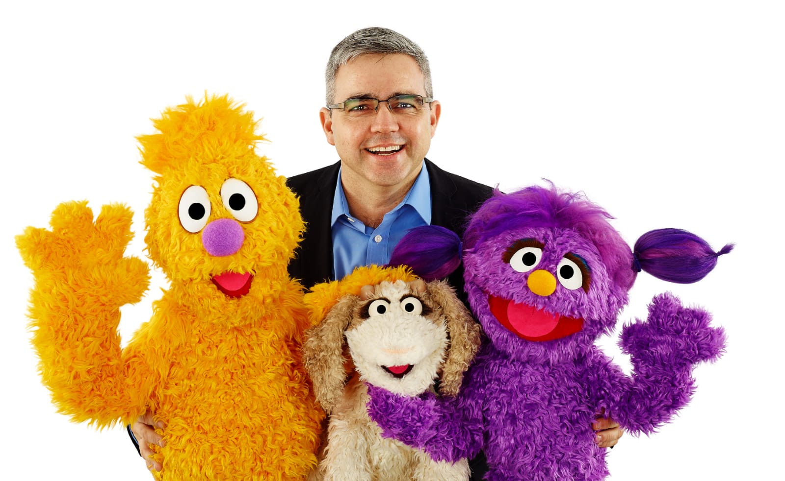 René Celaya, in a suit and glasses, smiles with three diverse puppets, reflecting global citizenship and commitment to education.