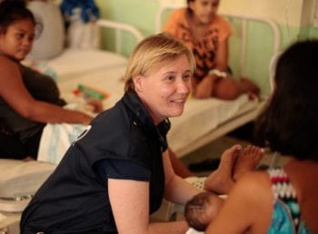 A diverse healthcare worker engages compassionately with a mother and baby, fostering global service and community well-being.